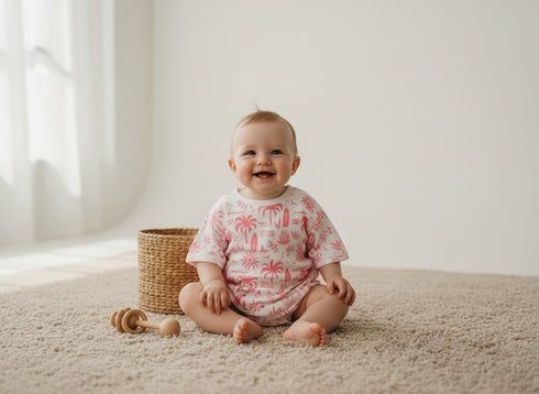 Baby sitting on a carpeted floor wearing a pink and white onesie with palm tree pattern, next to a wicker basket and wooden toy.