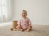 Baby sitting on a carpeted floor wearing a pink and white onesie with palm tree pattern, next to a wicker basket and wooden toy.