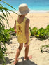 A young child wearing a yellow crochet romper and a beige bucket hat, photographed from behind on a sandy beach path with green plants and the ocean in the background.
