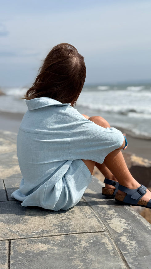 Person sitting on a stone path by the ocean wearing a light blue shirt.