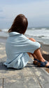 Person sitting on a stone path by the ocean wearing a light blue shirt.
