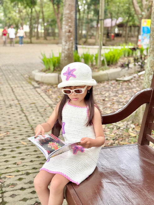 Young girl in a white dress and hat with a starfish design, sitting on a bench in a park.