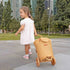Child in a white dress pushing a wicker toy cart in an urban park setting.