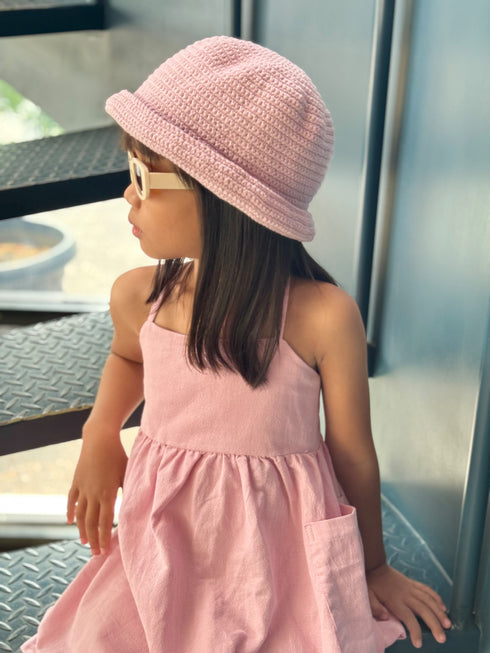 A young child wearing a light pink knit bucket hat and a matching pink sleeveless dress, photographed indoors near a window with metal steps visible in the background.