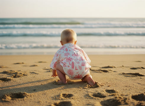 Baby in a floral outfit sitting on a sandy beach looking at the ocean.