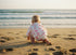 Baby in a floral outfit sitting on a sandy beach looking at the ocean.