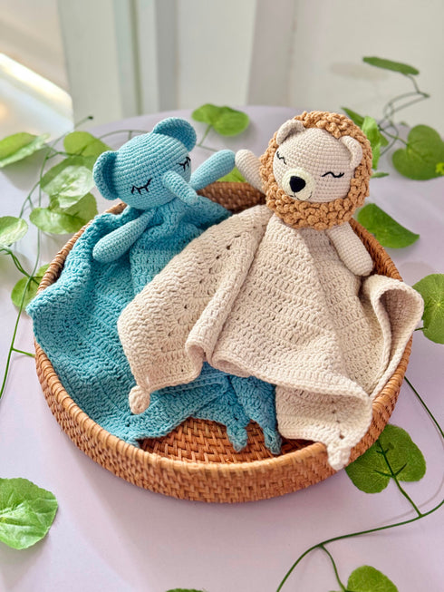 Two crocheted toys, a blue elephant and a beige lion, resting in a round woven basket on a light pink surface with green leafy vines.