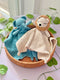 Two crocheted toys, a blue elephant and a beige lion, resting in a round woven basket on a light pink surface with green leafy vines.