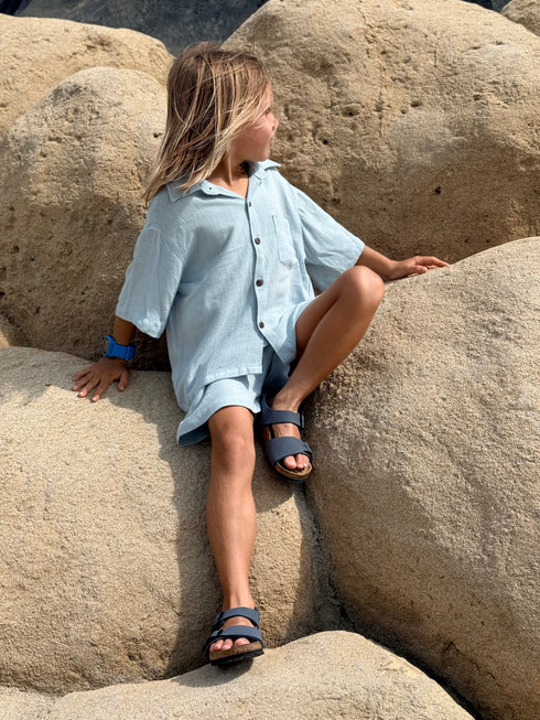 Child sitting on large rocks wearing a light blue shirt and dark sandals.