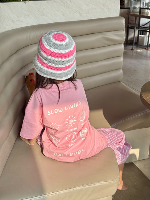 A child sitting on a beige leather bench, viewed from behind, wearing a pink t-shirt with 'SLOW LIVING' printed on the back, a pink and white striped knit hat, and a pink striped skirt.