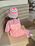 A child sitting on a beige leather bench, viewed from behind, wearing a pink t-shirt with 'SLOW LIVING' printed on the back, a pink and white striped knit hat, and a pink striped skirt.
