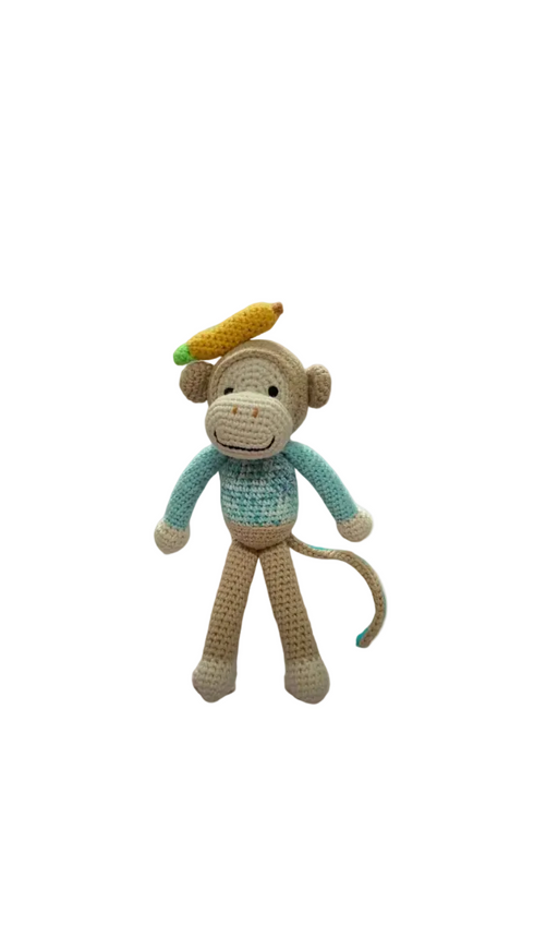 A crochet doll representing a red monkey with a blue shirt and a green accessory on its head, standing upright on a wooden surface.