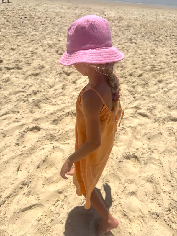 Child wearing a pink hat and yellow dress standing on sandy ground.