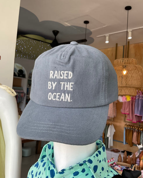 A gray cap with 'RAISED BY THE OCEAN' embroidered on the front, displayed on a mannequin mannequin in a retail setting.