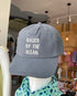A gray cap with 'RAISED BY THE OCEAN' embroidered on the front, displayed on a mannequin mannequin in a retail setting.