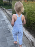 Child wearing a blue and white striped romper standing on a sidewalk with greenery in the background