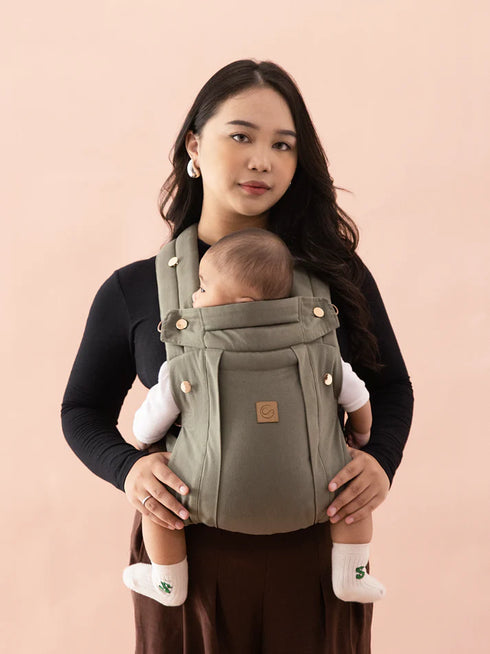 Woman carrying a baby in an olive green baby carrier against a pink background