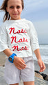 Child wearing a white shirt with 'Nasi Nasi Nasi' text by the ocean