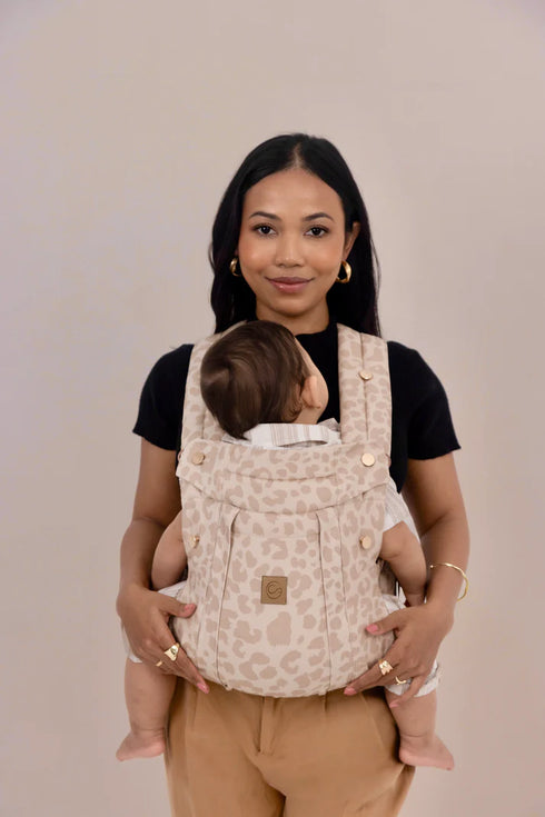 Woman holding a baby in a beige baby carrier against a plain background