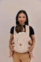 Woman holding a baby in a beige baby carrier against a plain background