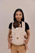 Woman holding a baby in a beige baby carrier against a plain background