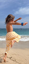 Person in yellow checkered shorts standing on one leg on a beach with ocean and sky in the background