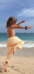 Person in yellow checkered shorts standing on one leg on a beach with ocean and sky in the background