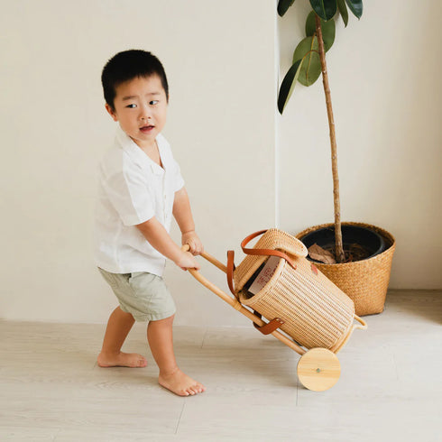 Child pushing a woven toy cart in a minimalistic room with a plant.