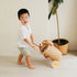 Child pushing a woven toy cart in a minimalistic room with a plant.