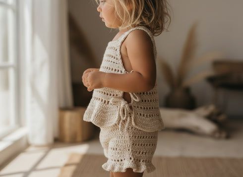 Child wearing a crochet outfit standing by a window in a softly lit room.