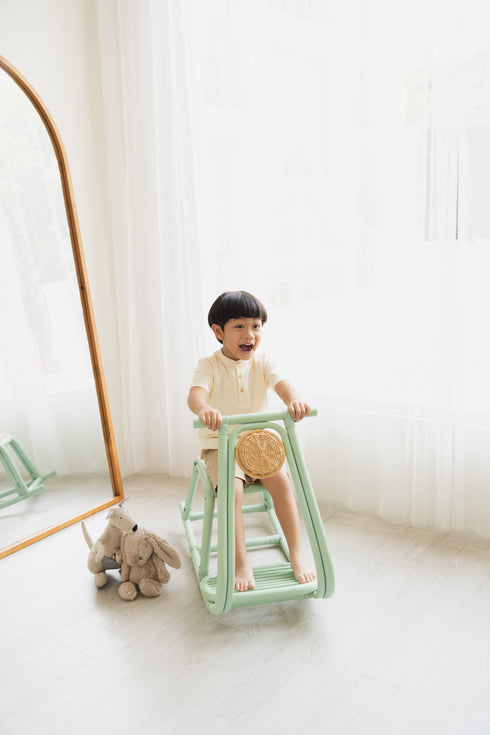 Child playing on a green toy in a room with white walls and a wooden floor.