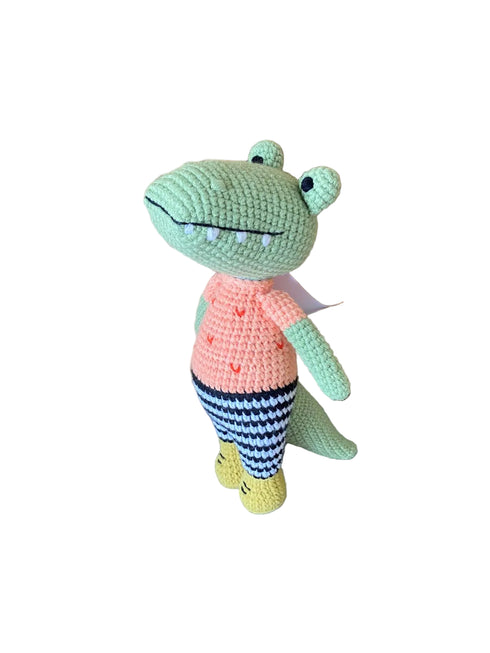 Crocheted frog toy wearing a peach shirt and striped pants on a wooden floor.