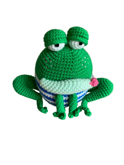Crocheted green frog toy with a striped shirt on a reflective surface