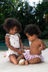 Two young children sitting on a tiled floor outdoors, surrounded by greenery.