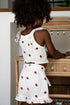 Child wearing a white dress with red strawberry patterns, standing in front of a wooden chair.