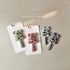 A set of three Koala Rattle Teethers in different colours packaged on white cardboard backing. Each teether features a bear-like design with a primary colour and a smaller secondary colour, and they all have a round rattle at the base.
