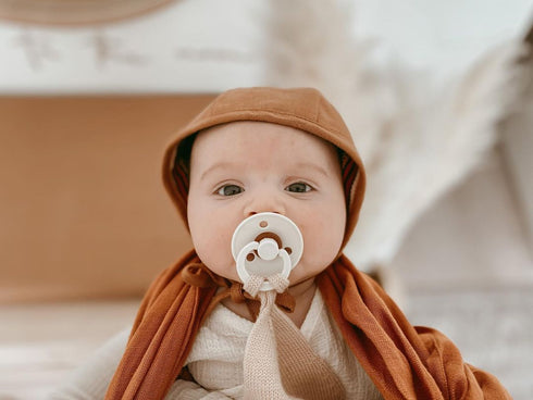 Baby in a rust bonnet with a BIBS Colour pacifier in Vanilla