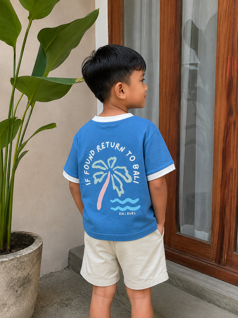 A young boy wearing a blue ringer t-shirt with a palm tree graphic and the text 'IF FOUND RETURN TO BALI' on the back, paired with beige shorts, standing in front of a wooden door.