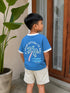 A young boy wearing a blue ringer t-shirt with a palm tree graphic and the text 'IF FOUND RETURN TO BALI' on the back, paired with beige shorts, standing in front of a wooden door.